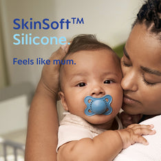 Baby with a blue MAM pacifier dummy held by a woman, with 'SkinSoft Silicone' text overlay.