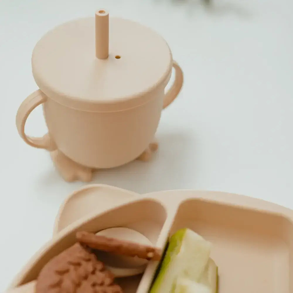 Beige sippy cup with a lid and straw, placed on a white surface with a beige plate containing cookies and a slice of fruit.