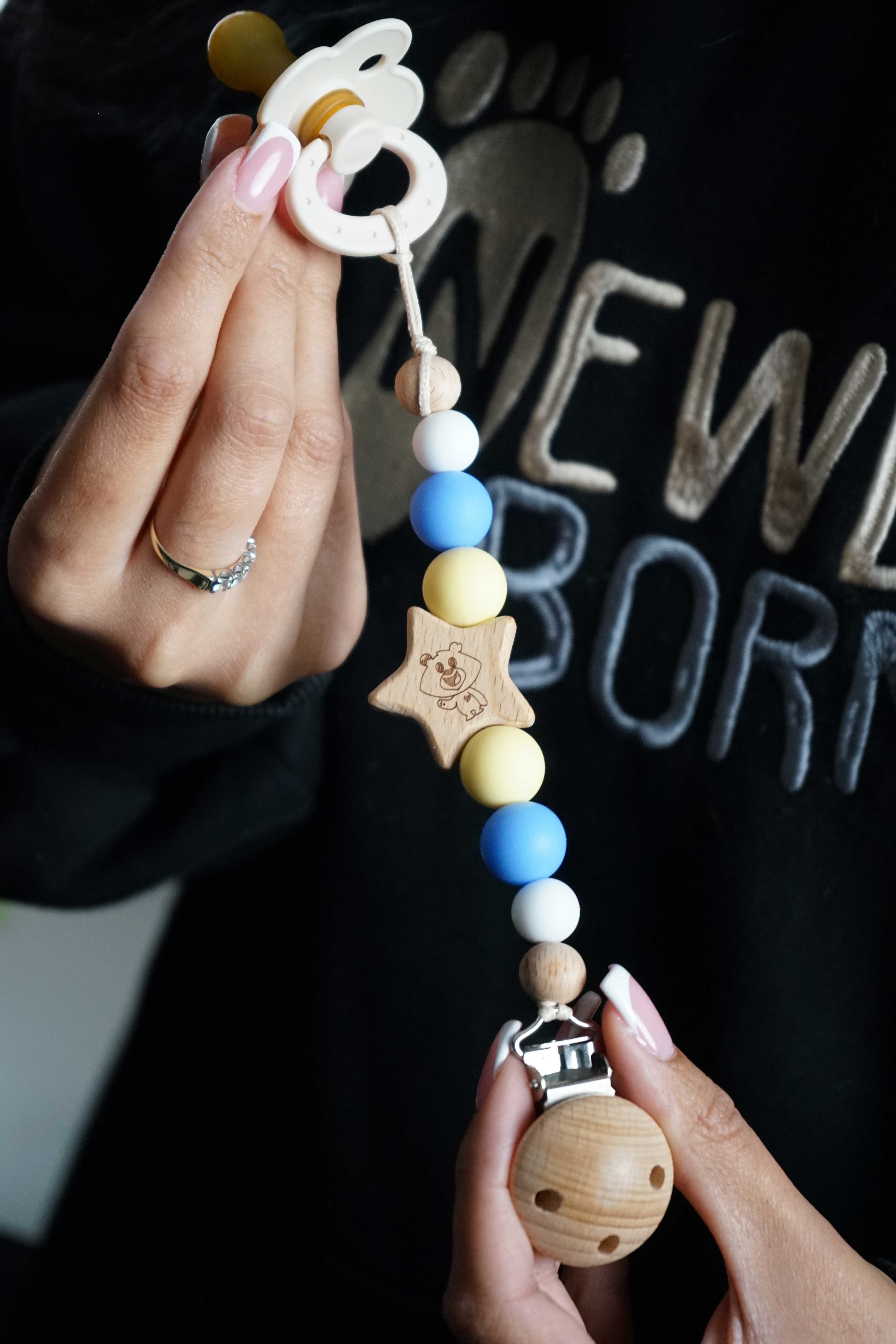 A baby pacifier clip and teether set held by Sasha's hands. The set features a white silicone pacifier, wooden beads in white, blue, yellow, and natural wood, a star-shaped wooden teething ring, and a natural wood ball clip. Collaboration with Sheffield Children's Hospital Charity