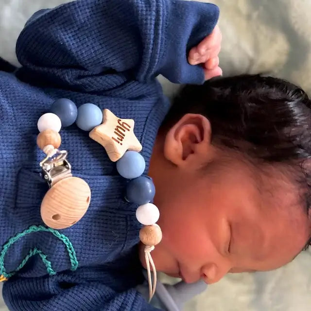 Newborn baby wearing a blue outfit with newly born personalised star dummy clip in blue, lying on a soft surface.