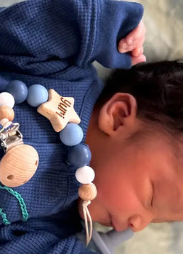 Newborn baby wearing a blue outfit with newly born personalised star dummy clip in blue, lying on a soft surface.