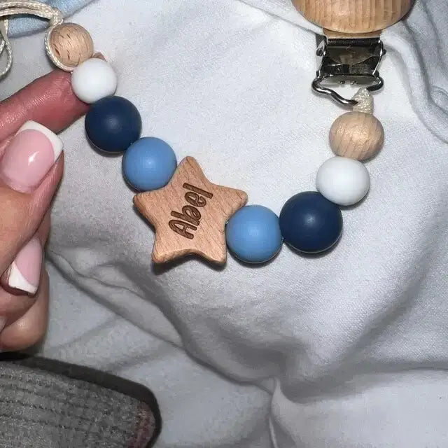 Silicone personaliesd pacifier clip with wooden star charm and 'Alex' engraving, held by a hand.