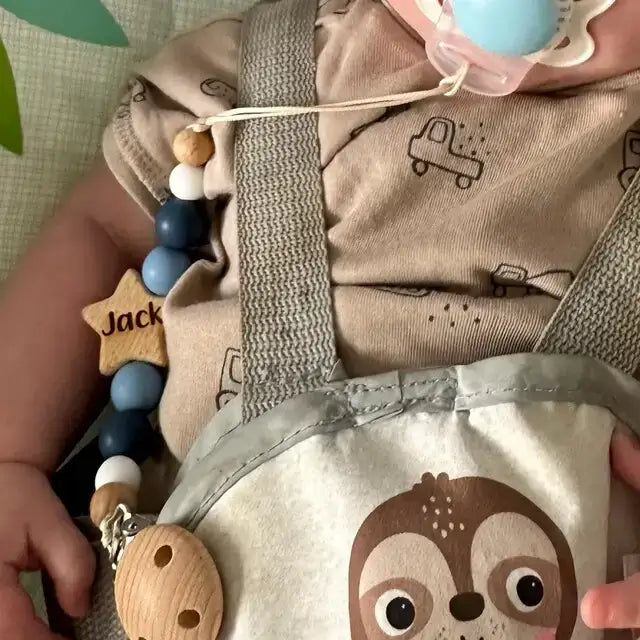 Baby holding a personalied star dummy clip in blue with 'Jack' on it, wearing a beige outfit with truck designs.