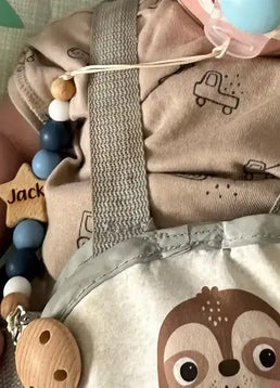 Baby holding a personalied star dummy clip in blue with 'Jack' on it, wearing a beige outfit with truck designs.
